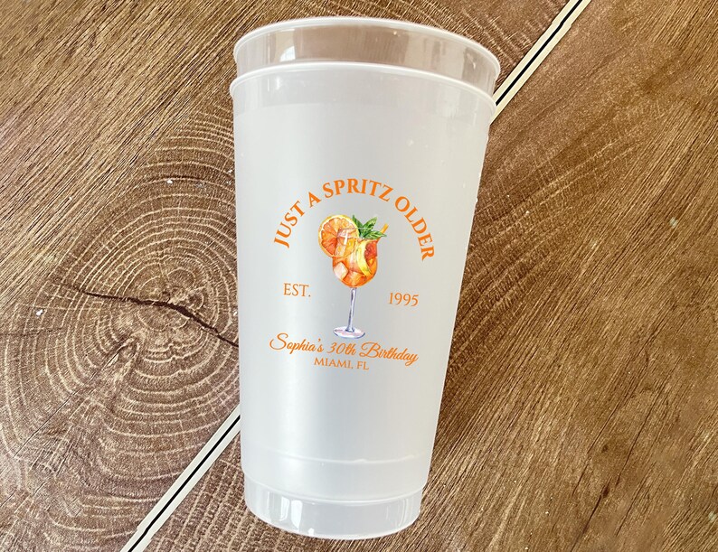 May include: A clear plastic cup with a frosted appearance. The cup features an orange graphic of a cocktail with the text "JUST A SPRITZ OLDER" and "EST. 1995". Below the graphic is the text "Sophia's 30th Birthday, Miami, FL".