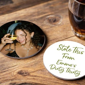 May include: Two round coasters on a wooden surface. One coaster features a photo of a woman holding a glass of wine. The other coaster has the text "Stole This From Emma's Dirty Thirty" in green script.