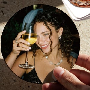 May include: A circular drinks coaster showing a person smiling and holding a glass of white wine. The person is wearing jewellery, including a pearl necklace and earrings. The coaster is held by a hand, with a second coaster and a glass of amber liquid in the background.