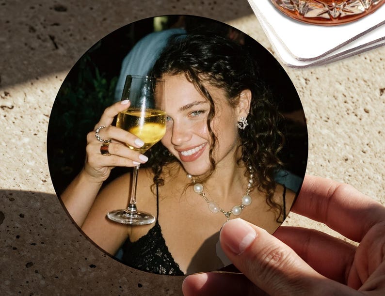 May include: A circular drinks coaster showing a person smiling and holding a glass of white wine. The person is wearing jewellery, including a pearl necklace and earrings. The coaster is held by a hand, with a second coaster and a glass of amber liquid in the background.