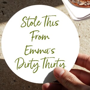 May include: A white, circular drinks coaster with green text. The text reads "Stole This From Emma's Dirty Thirty." The coaster is held by a hand, with a blurred background.