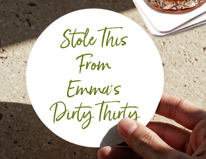 May include: A white, circular drinks coaster with green text. The text reads "Stole This From Emma's Dirty Thirty." The coaster is held by a hand, with a blurred background.