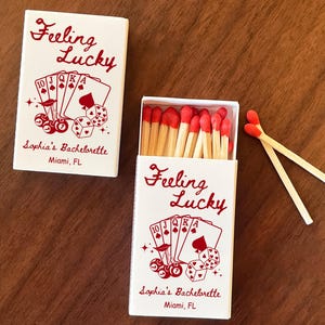 May include: Two white matchboxes with red text and graphics. One box is closed, the other open, revealing red-tipped matches. The text reads "Feeling Lucky" and "Sophia's Bachelorette, Miami, FL". The design includes playing cards and dice.