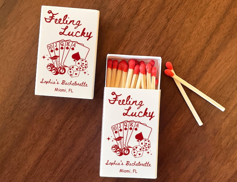 May include: Two white matchboxes with red text and graphics. One box is closed, the other open, revealing red-tipped matches. The text reads "Feeling Lucky" and "Sophia's Bachelorette, Miami, FL". The design includes playing cards and dice.