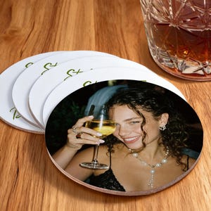 May include: White circular coasters featuring a photograph of a woman with a glass of wine. The coasters have a green script design. A glass of amber liquid is nearby on a wooden surface.