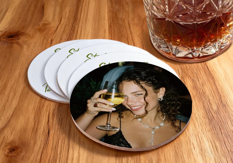 May include: White circular coasters featuring a photograph of a woman with a glass of wine. The coasters have a green script design. A glass of amber liquid is nearby on a wooden surface.