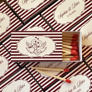 May include: A collection of matchboxes with a burgundy and white striped design. The matchbox in the center features a swan illustration and the names "Sophia & Peter" with the date "11.06.2026". The matches have red tips.