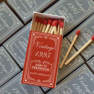 May include: A collection of matchboxes with a vintage design. The open red matchbox displays wooden matches with red tips. The boxes are gray with white text that reads "Vintage 1995" and "Aged to Perfection Happy Birthday!"