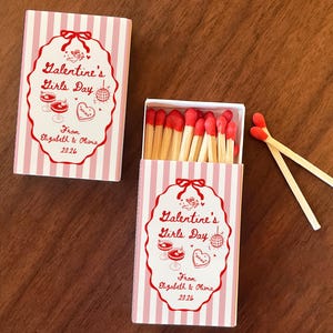 May include: Two matchboxes with a pink and white striped design. The boxes feature a red oval label with the text "Galentine's Girls Day" and the names "Elizabeth & Olivia 2026". Several matches are visible inside one box, and two matches are outside.
