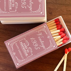 May include: A set of matchboxes with a vintage design. The boxes are a dusty rose color with white text that reads "Vintage 1995" and "Aged to Perfection, Happy Birthday!". The matches have red tips.