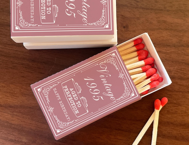 May include: A set of matchboxes with a vintage design. The boxes are a dusty rose color with white text that reads "Vintage 1995" and "Aged to Perfection, Happy Birthday!". The matches have red tips.