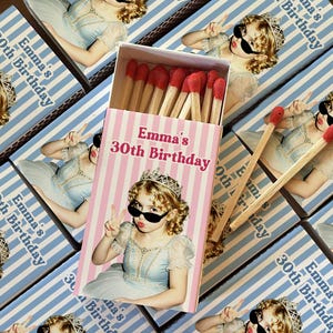May include: A rectangular matchbox with a pink and white striped cover featuring the text Emma's 30th Birthday and a photograph of a person wearing a tiara and sunglasses. The box is filled with wooden matches that have red tips.