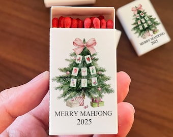May include: A white matchbox featuring a Christmas tree design made of Mahjong tiles, topped with a pink bow. The text "MERRY MAHJONG 2025" is printed below the tree. The match heads are red.