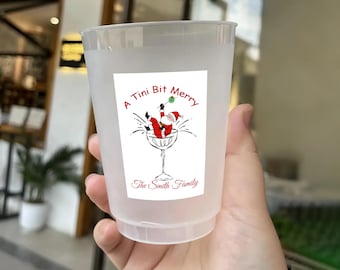 May include: A clear plastic cup with a festive holiday design. The design features Santa Claus in a martini glass with the text "A Tini Bit Merry" and "The Smith Family" in red. The cup is held in a hand.
