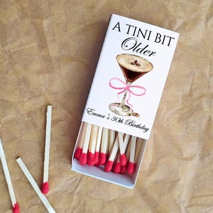 May include: A white matchbox with the text "A TINI BIT Older" and a watercolor illustration of a martini glass with a pink bow. The box is open, revealing red-tipped matches. The text "Emma's 30th Birthday" is also visible.