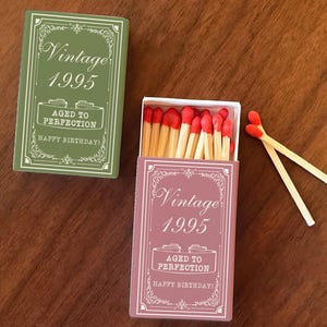May include: Two boxes of matches, one green and one pink, with the text "Vintage 1995" and "Aged to Perfection" printed on them. The boxes are open, revealing red-tipped matches. Two matches are lying outside the pink box.