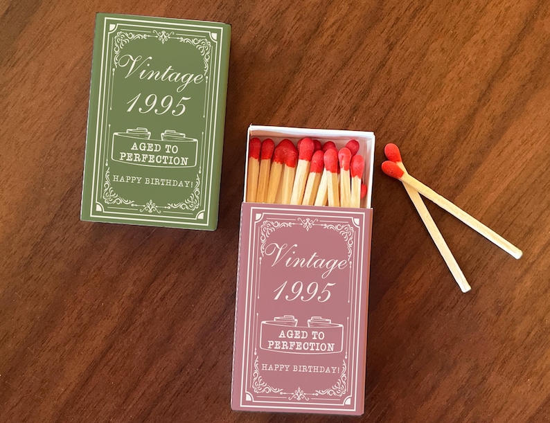 May include: Two boxes of matches, one green and one pink, with the text "Vintage 1995" and "Aged to Perfection" printed on them. The boxes are open, revealing red-tipped matches. Two matches are lying outside the pink box.