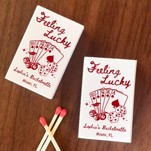 May include: Two white matchboxes with red text that reads "Feeling Lucky" and "Sophia's Bachelorette, Miami, FL." The boxes feature a poker-themed design with playing cards, dice, and cherries. Three matches with red tips are also visible.