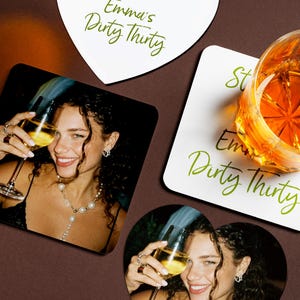 May include: Coasters featuring a photograph of a smiling person holding a glass of wine. The coasters display the text "Emma's Dirty Thirty" in green script. A glass of amber liquid sits on a coaster. The coasters are square, heart, and round.