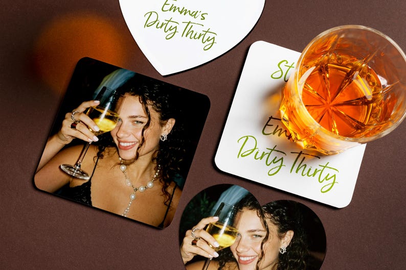 May include: Coasters featuring a photograph of a smiling person holding a glass of wine. The coasters display the text "Emma's Dirty Thirty" in green script. A glass of amber liquid sits on a coaster. The coasters are square, heart, and round.