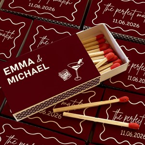 May include: Burgundy matchboxes with white text that reads "EMMA & MICHAEL" and "the perfect match 11.06.2026". The matchbox is open, revealing wooden matches with red tips. A cocktail graphic is also visible.