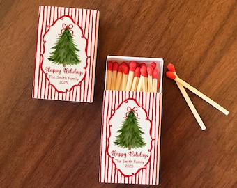 May include: Two matchboxes with a red and white striped design. Each box features a watercolor Christmas tree illustration with the text "Happy Holidays The Smith Family 2025". Two matches are next to the open box.