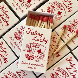 May include: White matchboxes with red text and illustrations. The boxes feature the words "Feeling Lucky" and a design of playing cards and dice. The text "Sophia's Bachelorette Miami, FL" is also printed on the boxes.