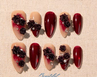 Può includere: Set di dieci unghie finte. Le unghie presentano una combinazione di design, tra cui bordeaux pieno e unghie color nude con accenti bordeaux e abbellimenti di rose nere. Alcune unghie hanno accenti di perle e strass. La parola "Beautiful" è scritta in basso.