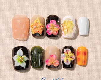 May include: A collection of ten artificial nails with 3D floral designs. The nails feature various colors, including pink, yellow, white, and orange, with a glossy finish. Some nails have a clear, droplet-like texture.