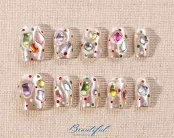 Peut inclure: Dix faux ongles beige avec des strass et des paillettes colorés. Les ongles sont disposés en deux rangées de cinq. Le texte "Beautiful Handmade Nail" est en bas de l'image.