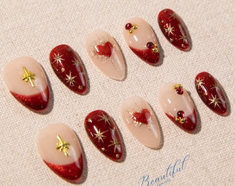 May include: A set of ten almond-shaped artificial nails. The nails feature a variety of designs, including a deep red glitter base with gold star accents, a nude base with a red heart design, and a nude base with a gold star or jewel accents. The word "Beautiful" is written on the bottom right.