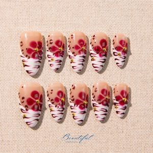 May include: A set of ten artificial nails with a floral design. The nails are almond-shaped with a nude base, adorned with red floral patterns, gold accents, and a white and brown zebra stripe design at the tips. The word "Beautiful" is written in cursive.