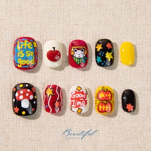 May include: A collection of ten press-on nails with diverse designs. The nails showcase vibrant artwork, including the text "life is so good" and "Good Time", a red apple, a cat in a hat, stars, and a mushroom. The nails are displayed on a neutral backdrop.