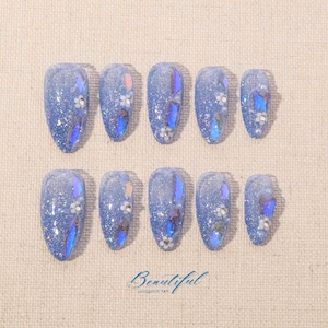 Puede incluir: Conjunto de diez uñas postizas azules e iridiscentes con detalles de purpurina y flores. Las uñas están dispuestas en dos filas de cinco. El texto "Beautiful Handmade Nail" está impreso debajo de las uñas.