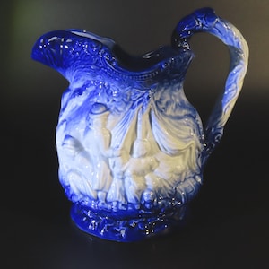 May include: A blue and white ceramic jug with a detailed relief design. The jug features a curved handle and a spout. The relief depicts figures and foliage. The jug is a decorative item.