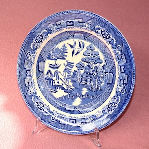 Può includere: Un piatto rotondo blu e bianco con motivo Willow. Il piatto presenta una scena paesaggistica dettagliata con alberi, edifici e uccelli. Il bordo ha un motivo geometrico ripetitivo. Il piatto è esposto su un supporto trasparente.