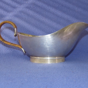 Puede incluir: Una salsera plateada con asa trenzada. El recipiente tiene un pico curvo y una banda dorada decorativa alrededor de la base. El asa es de material tejido marrón claro.