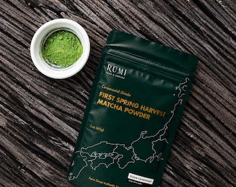 May include: A dark green pouch of Rumi Teas & Coffee First Spring Harvest Matcha Powder, 2oz (60g), featuring a white map outline and text. Beside it sits a small white bowl containing a portion of vibrant green matcha powder.