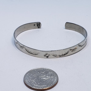 May include: Silver-toned cuff bracelet with etched Southwestern-style designs, including sun and mountain motifs. A U.S. quarter coin is next to the bracelet for size comparison. The bracelet is on a white surface.