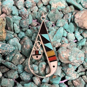 May include: A turquoise, black, red, yellow, and white geometric pendant with a silver bail. The pendant is shaped like a teardrop and has a small circle in the center.