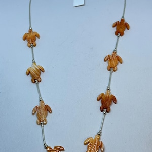 Könnte beinhalten: Eine silberne Halskette mit mehreren orangefarbenen Schildkrötenanhängern. Die Schildkröten sind auf einer dünnen Silberkette mit kleinen Silberperlen zwischen jeder Schildkröte aufgereiht. Die Halskette hat einen Verschluss.