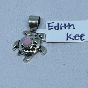 Könnte beinhalten: Ein silberner Schildkrötenanhänger mit einem rosa Mittelstein. Der Anhänger hat eine Schlaufe für eine Kette und liegt auf einer hellgrauen Oberfläche. Das Etikett daneben lautet "Edith Kee".