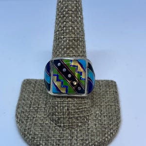 Puede incluir: Un anillo de plata con una cara rectangular con un diseño geométrico colorido. El diseño incluye secciones azules, verdes, amarillas y negras con pequeños puntos blancos. El anillo se muestra en un soporte para anillos texturizado de color neutro.