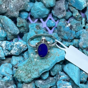 Op de afbeelding: Een zilveren ring met een donkerblauwe ovale steen, gepresenteerd op een bed van turkooizen stenen. De ring heeft decoratieve elementen rond de steen. De turkooizen stenen zijn van verschillende groottes en tinten.