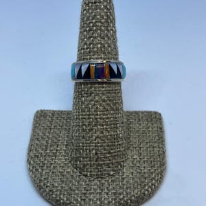 Peut inclure: Une bague en argent avec une bande ornée de motifs géométriques incrustés de couleurs turquoise, noir, blanc, orange et irisée. La bague est présentée sur un présentoir à bagues texturé et beige, sur fond blanc.