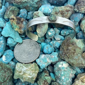 May include: A silver cuff bracelet with a turquoise stone set in the center. The bracelet has etched details and is displayed on a bed of turquoise stones. A quarter is next to the bracelet for size reference.