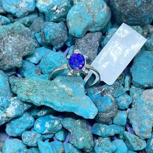 May include: A silver ring with a blue opal stone in a turtle design. The ring is set against a background of turquoise stones. The opal has a vibrant, iridescent quality.
