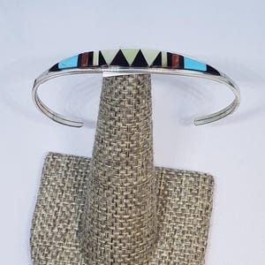 Op de afbeelding: Een zilveren manchetarmband met een geometrisch inlegontwerp. De inleg heeft turquoise, zwarte, witte en bruine segmenten. De armband wordt gepresenteerd op een bruine stoffen sieradenstandaard.