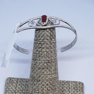 Puede incluir: Un brazalete plateado con una piedra roja ovalada en el centro. El brazalete presenta remolinos decorativos a ambos lados de la piedra. La joya se exhibe sobre una superficie texturizada de color neutro.