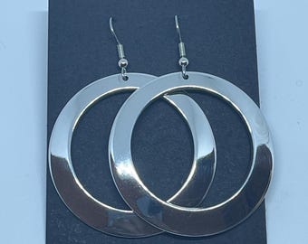 Puede incluir: Un par de pendientes de aro en tono plateado pulido con un diseño circular ancho y plano. Los pendientes cuelgan de ganchos de metal plateado y se presentan sobre una tarjeta rectangular de color negro liso.